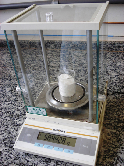 Béquer sendo pesado numa balança analítica, que mostra 58,4428 g