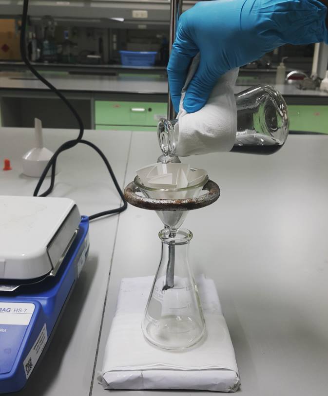 Líquido sendo despejado num funil de vidro com papel filtro, sendo escoado num frasco de vidro