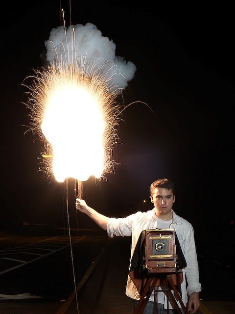Homem, atrás de uma câmera fotográfica antiga, segurando um bastão com uma grande explosão na ponta, de cor amarelada e soltando faíscas