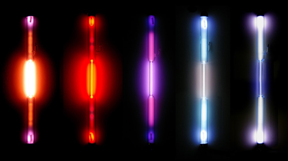 Cinco lâmpadas cilíndricas, lado a lado, brilhando com as seguintes cores, da esquerda para a direita: laranja, vermelha, roxa, azul clara, roxa clara