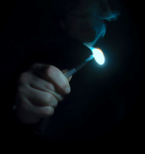 Uma mão segurando uma pinça metálica, com algo queimando na ponta, mostrando uma luz branca e soltando fumaça